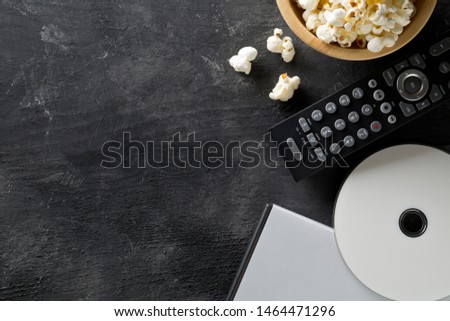 DVD or blu ray movie disc with tv remote control and bowl of popcorn on dark background. Home theatre movie or series night concept. Flat lay top view from above with copy space.