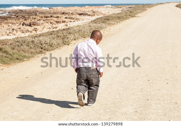 dwarfish african man walking down a dirt road\
along the coast with his head bent\
down