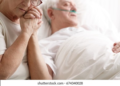Dying senior man in hospital bed and his sad wife holding his hand