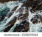 Dynamic aerial view of powerful ocean waves crashing onto the rugged, rocky coastline, surrounded by swirling seafoam, capturing the untamed energy and natural beauty of a scenic seaside environment.