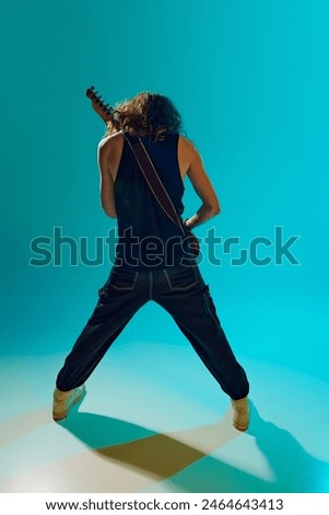 Dynamic and artistic performance of young man with long hair playing electric guitar against blue, cyan background in neon light. Concept of music, performance, festival, live concert