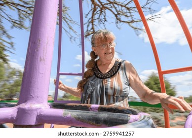 A dynamic and authentic medium shot of a senior woman with visible burn scars on her face and hands, joyfully spinning on a colorful metal roundabout at an outdoor playground. The motion blur of the trees and the bright blue sky in the background emphasize a sense of energy, freedom, and an active lifestyle