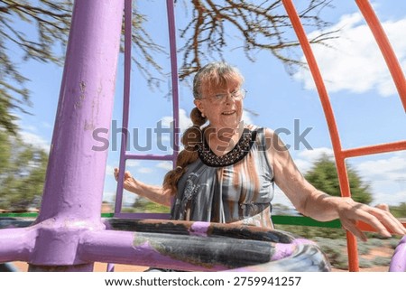 A dynamic and authentic medium shot of a senior woman with visible burn scars on her face and hands, joyfully spinning on a colorful metal roundabout at an outdoor playground. The motion blur of the trees and the bright blue sky in the background emphasize a sense of energy, freedom, and an active lifestyle