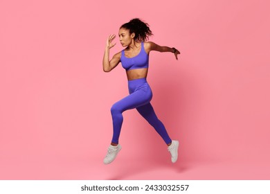 Dynamic fitness. Motivated black sportswoman in purple sportswear leaps and runs in mid air, showcasing strength and athleticism over pink studio background, full length shot - Powered by Shutterstock