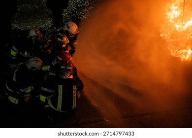 A dynamic, high-angle close-up shot of a huddle of firefighters wearing helmets (some with red and white colors) as they direct a powerful stream of water. The stream fills the right side of the frame, backlit by the massive orange fire, creating a strong contrast between the dark figures and the bright light.