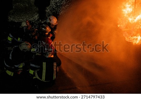 A dynamic, high-angle close-up shot of a huddle of firefighters wearing helmets (some with red and white colors) as they direct a powerful stream of water. The stream fills the right side of the frame, backlit by the massive orange fire, creating a strong contrast between the dark figures and the bright light.