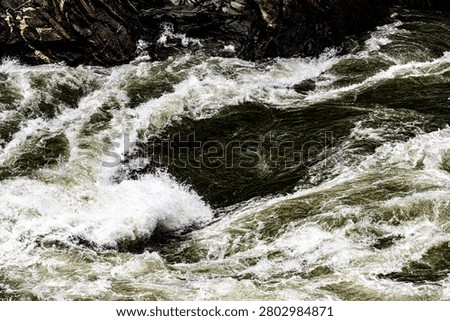 Dynamic high-angle shot capturing the fierce convergence of raging river currents in the Jinsha River inside Tiger Leaping Gorge, Yunnan Province, China. Heavy, dark olive-green waters sweep diagonally across the frame, exploding into a turbulent wake of bright white foam as they collide with a submerged rock shelf. The wet, craggy slate visible at the upper left provides a solid geological boundary to the unstoppable kinetic drive of this world-famous wild waterway.