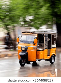 A dynamic image captures a bright orange auto rickshaw in motion on a rain-slicked city street. The rickshaw, with its dark brown top and patterned interior, is sharply focused against a blurred 