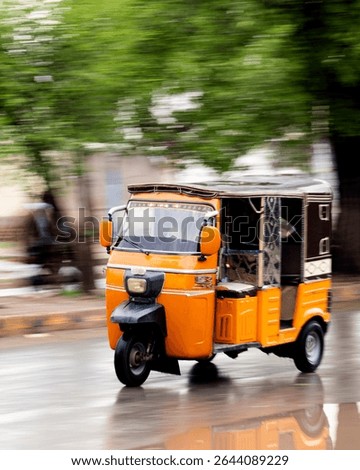 A dynamic image captures a bright orange auto rickshaw in motion on a rain-slicked city street. The rickshaw, with its dark brown top and patterned interior, is sharply focused against a blurred 