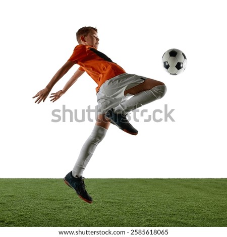 Dynamic image of focused boy in orange jersey and white shorts in motion, kicking ball with knee, training against white background. Concept of sport, education, competition, game