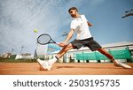 Dynamic image of focused young man in white t-shirt playing tennis on outdoor clay court, showing energy and focus during match. Concept of sport, competition, active and healthy lifestyle