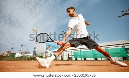Dynamic image of focused young man in white t-shirt playing tennis on outdoor clay court, showing energy and focus during match. Concept of sport, competition, active and healthy lifestyle