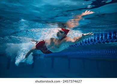 Dynamic image of young guy, swimming athlete in red cap and goggles in motion in swimming pool, training. Freestyle swim. Concept of professional sport, health, endurance, strength, active lifestyle