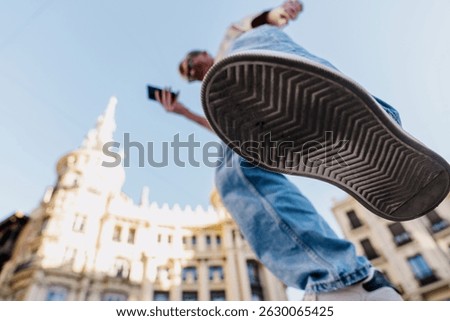 Dynamic low angle shot of stylish young man holding smartphone while walking in sunny European city, emphasizing freedom, movement and active urban lifestyle with historic buildings in background