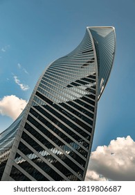 Dynamic low angle shot of a twisted glass skyscraper facade against a bright blue sky with clouds. High-tech architecture and urban business concept.