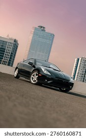Dynamic low angle view of a black sports coupe parked on a rooftop during golden hour with modern city skyscrapers and purple sunset sky.