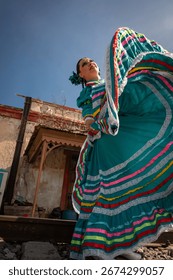 Dynamic low angle view of a Mexican woman performing a traditional folkloric dance in a turquoise dress with multicolor ribbons, set against urban architecture and blue sky.