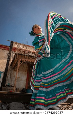 Dynamic low angle view of a Mexican woman performing a traditional folkloric dance in a turquoise dress with multicolor ribbons, set against urban architecture and blue sky.