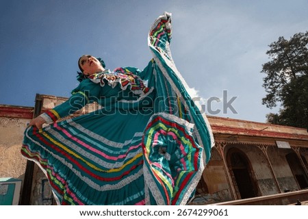 Dynamic low angle view of a Mexican woman performing a traditional folkloric dance in a turquoise dress with multicolor ribbons, set against urban architecture and blue sky.