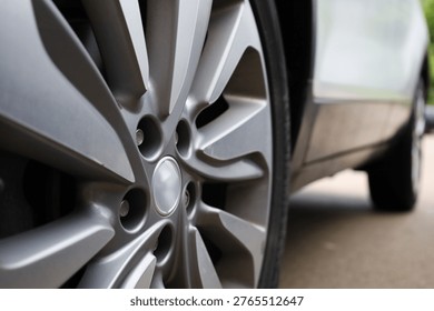 A dynamic, low-angle close-up of a multi-spoke alloy car wheel. The shot features a shallow depth of field, bringing the matte silver finish of the rim and the center cap into sharp focus while the tire and the side of the white vehicle softly blur into the background. The composition emphasizes the sleek lines, lug nut wells, and the metallic texture of the wheel's design.