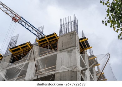 Dynamic low-angle perspective of multi-story building under construction, featuring raw concrete, exposed rebar, temporary yellow formwork, protective safety nets, and prominent tower crane against ov