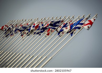 A dynamic low-angle shot captures numerous Georgian, European Union, and Greek flags flying vigorously from an array of slanted white poles. The vibrant flags flutter against a bright, clear sky, symbolizing international relations and cultural diversity in Batumi, Georgia.
