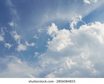  A dynamic low-angle shot of a vibrant blue sky featuring fluffy white cumulus clouds on the right, accompanied by subtle sunbeams filtering through soft wispy cirrus cloud textures