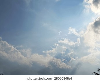  A dynamic low-angle shot of a vibrant blue sky featuring fluffy white cumulus clouds on the right, accompanied by subtle sunbeams filtering through soft wispy cirrus cloud textures
