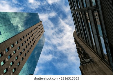A dynamic, low-angle upward perspective looking between modern urban skyscrapers toward a vibrant blue sky filled with wispy white clouds. On the left, a contemporary glass-paneled high-rise reflects the sky, while an older commercial structure frames the right side. This composition highlights architectural contrast, urban density, corporate growth, and modern city design.