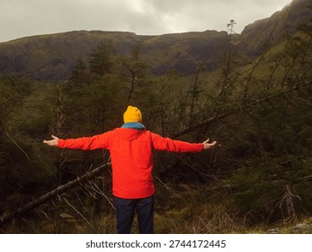 Dynamic man in a red jacket stands in a forest with his arms high. He is wearing a yellow hat and sunglasses. Scene is calm and peaceful, as the man is surrounded by nature. Travel and tourism
