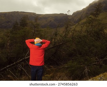 Dynamic man in a red jacket stands in a forest with his arms high. He is wearing a yellow hat and sunglasses. Scene is calm and peaceful, as the man is surrounded by nature. Travel and tourism