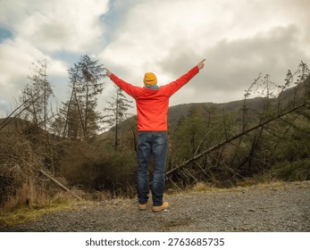 Dynamic man in a red jacket stands in a forest with his arms high. He is wearing a yellow hat and sunglasses. Scene is calm and peaceful, as the man is surrounded by nature. Travel and tourism