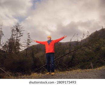 Dynamic man in a red jacket stands in a forest with his arms high. He is wearing a yellow hat and sunglasses. Scene is calm and peaceful, as the man is surrounded by nature. Travel and tourism