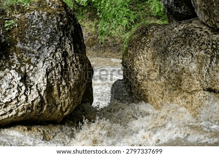 A dynamic medium shot capturing a powerful hydrological event inside a rocky mountain canyon. Two massive, weathered limestone boulders create a natural river obstruction and narrow bottleneck, forming a turbulent waterfall cascade. During a severe seasonal flash flood, the mud-brown, silt-laden water of the Sakhray river violently accelerates through the rocky gap, generating heavy white spray and churning foam. The rugged rock textures display intense chemical weathering and karstic erosion, typical of the Western Caucasus geomorphology in Adygea. This horizontal composition focuses on natural dams, riverbed blockages, and hydraulic dynamics, serving as an exceptional visual asset for hydrology textbooks, geomorphology journals, and environmental risk reporting