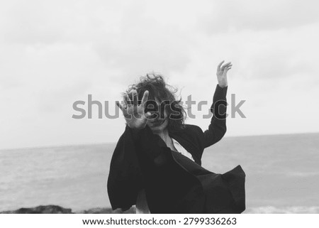 Dynamic movement in a wind swept scene, a flowing coat and raised arms against a seascape, conveying energy, freedom, and motion on a gray beach.
