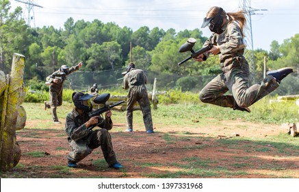 Dynamic paintball battle. Portrait female player jumping and aiming marker on member of opposing team