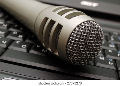 Dynamic performance technology: classic microphone positioned prominently on glossy computer keyboard, blending audio apparatus with modern digital interface. Sleek matte grey finish and distinctive metal mesh grille showcase robust design with intricate craftsmanship, suitable for both studio settings and live productions. Shot angle enhances dimensionality as microphone rests partially on keyboard's black keys, emphasizing technological synergy between sound equipment and computing hardware. Soft bokeh effect blurs keyboard background, directing viewer focus onto focal point while natural diffused lighting casts gentle reflections on metallic surface. Subdued grey and black color palette creates professional atmosphere resonating with themes of creativity, broadcasting, and artistic expression. Tight frame composition with subtle perspective contributes to intimate narrative about technological artistry and audio recording essentials, perfect for media production industries.