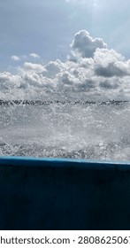 A dynamic point-of-view (POV) perspective from a moving boat, capturing an exhilarating spray of seawater exploding from the side. The image features a blue boat gunwale in the foreground, which frames the powerful, frozen-in-motion splash of water. In the background, a beautiful turquoise ocean expands under a vast blue sky filled with dramatic, fluffy white cumulus clouds, creating a refreshing and adventurous atmosphere for travel and marine content. Perfect for illustrating travel, adventure,