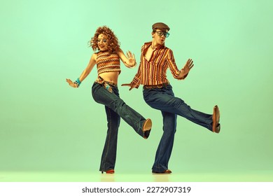 Dynamic portrait of happy man and woman in retro colored shirts and flared jeans dancing energetic dance over green background. Concept of fashion trends of 70s, 1980s years, music, hippie lifestyle