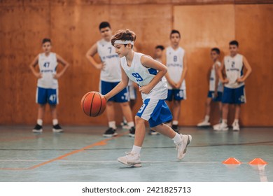 Dynamic portrait of a junior basketball player in action dribbling a ball and practicing basketball moves on court during the training.In blurry background are his teammates watching him.Boy with ball - Powered by Shutterstock