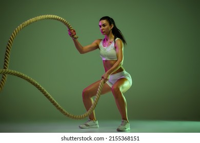 Dynamic Portrait Of Young Sportive Woman Training, Doing Exercises With Rope Isolated Over Green Studio Background In Neon Light. Concept Of Sport, Fitness, Healthy Lifestyle, Strength, Youth, Ad