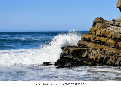 Dynamic scene of a large, powerful ocean wave crashing into stratified coastal rocks beneath a bright, clear blue sky. Features water splash and foam.