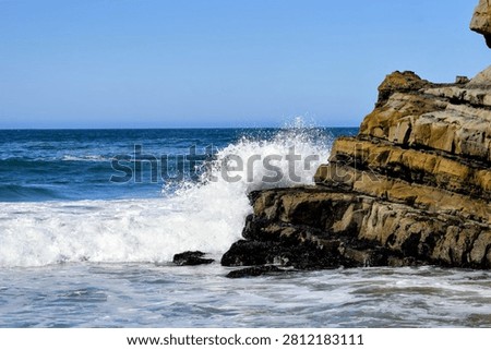 Dynamic scene of a large, powerful ocean wave crashing into stratified coastal rocks beneath a bright, clear blue sky. Features water splash and foam.