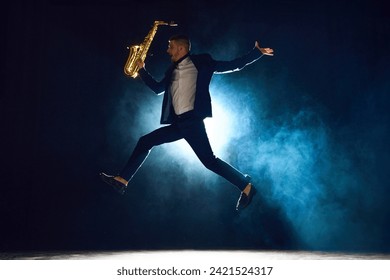 Dynamic shot of artistic man, solo performer jumping while playing saxophone on stage with backlights against dark background with smoke. Concept of art, instrumental music, dance, culture. Ad