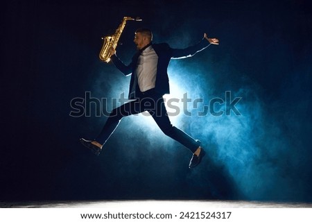 Dynamic shot of artistic man, solo performer jumping while playing saxophone on stage with backlights against dark background with smoke. Concept of art, instrumental music, dance, culture. Ad