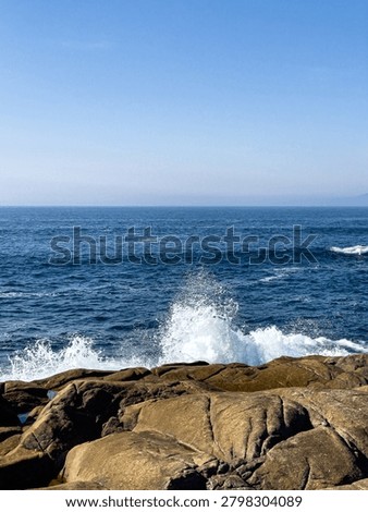 A dynamic shot of a white ocean wave splashing dramatically against smooth, sunlit granite rocks on the coastline under a clear blue sky.