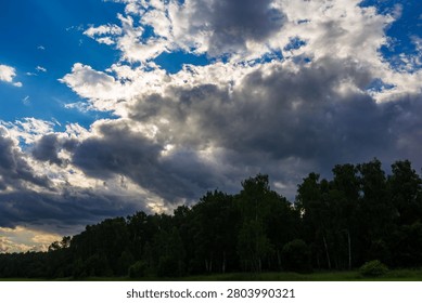 Dynamic sky with brewing storm clouds, vibrant blue patches, and sun rays breaking through. Dark, lush forest line below, adding depth to the dramatic scene