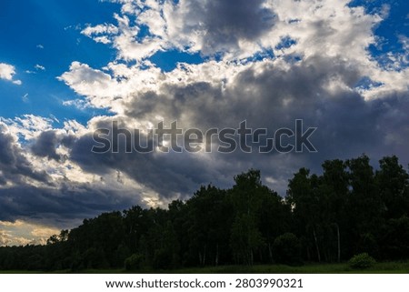 Dynamic sky with brewing storm clouds, vibrant blue patches, and sun rays breaking through. Dark, lush forest line below, adding depth to the dramatic scene