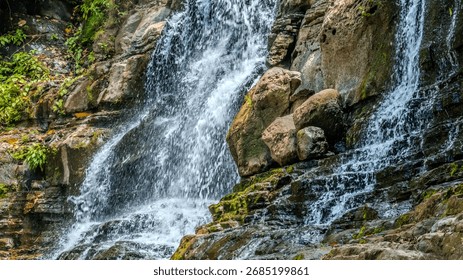 A dynamic, split waterfall rushes over rugged, moss-covered rock formations and large, smooth boulders. Lush green foliage frames the beautiful, powerful flow of nature.