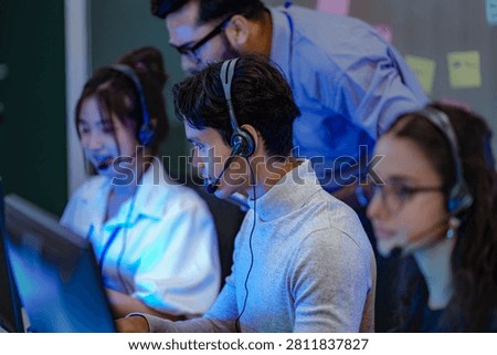 A dynamic team of tech support agents wearing headsets and operating desktop computers while a supervisor monitors network performance logs behind them.